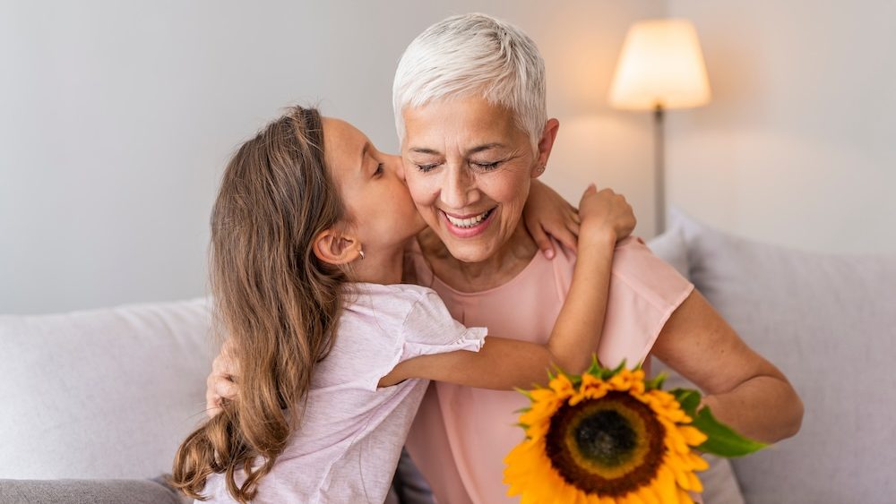 Little preschool granddaughter kissing happy older grandma on cheek giving sunflower congratulating smiling senior grandmother with birthday, celebrating mothers day or 8 march concept